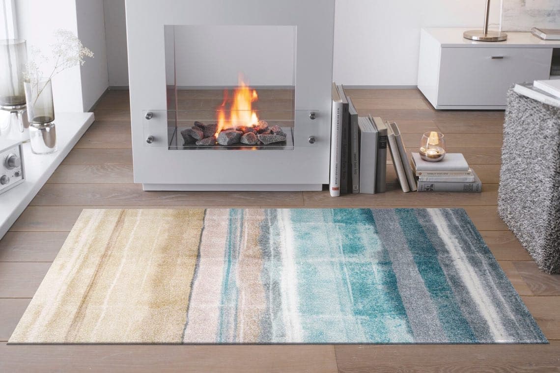 Moderner Kamin mit brennendem Feuer, daneben Bücher und eine Kerze, bunter Teppich auf Holzboden.
