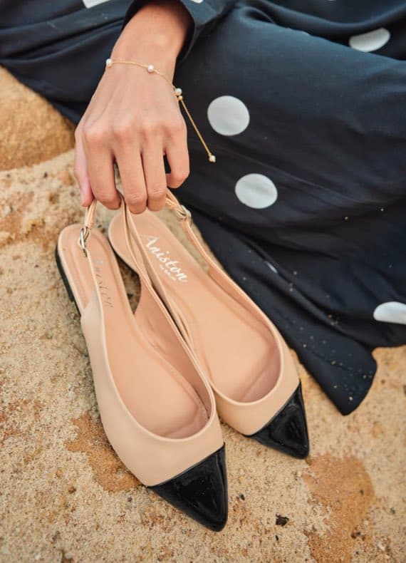 Frau hält beige Slingback-Schuhe mit schwarzer Spitze, trägt ein gepunktetes Kleid und ein Perlenarmband.