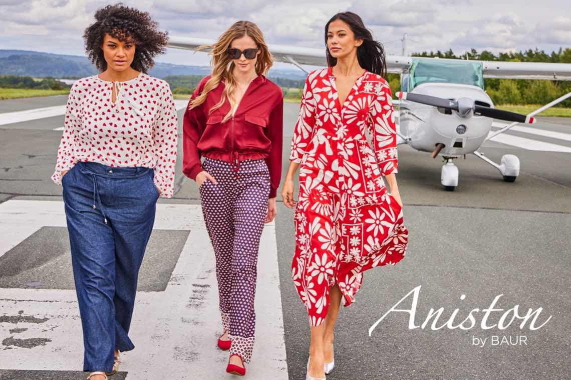 Aniston by BAUR – Drei Frauen in gemusterten Outfits laufen vor einem Flugzeug auf einer Landebahn.