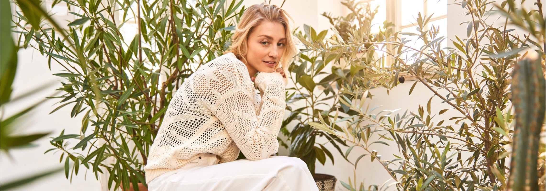 Person in hellem Strickpullover sitzt zwischen großen grünen Zimmerpflanzen in einem sonnigen Raum