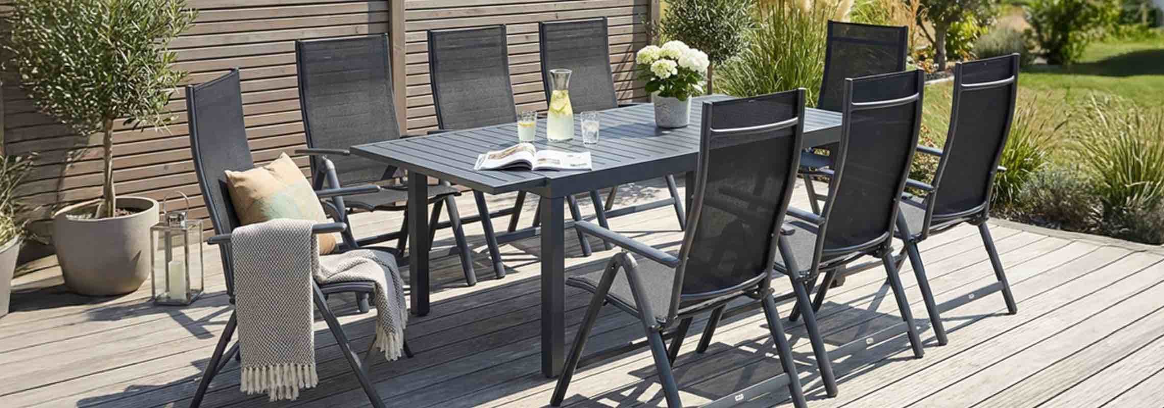 Große Gartentafel mit acht Stühlen auf Holzterrasse, darauf Karaffe, Gläser, Buch und Blumen, umgeben von Pflanzen