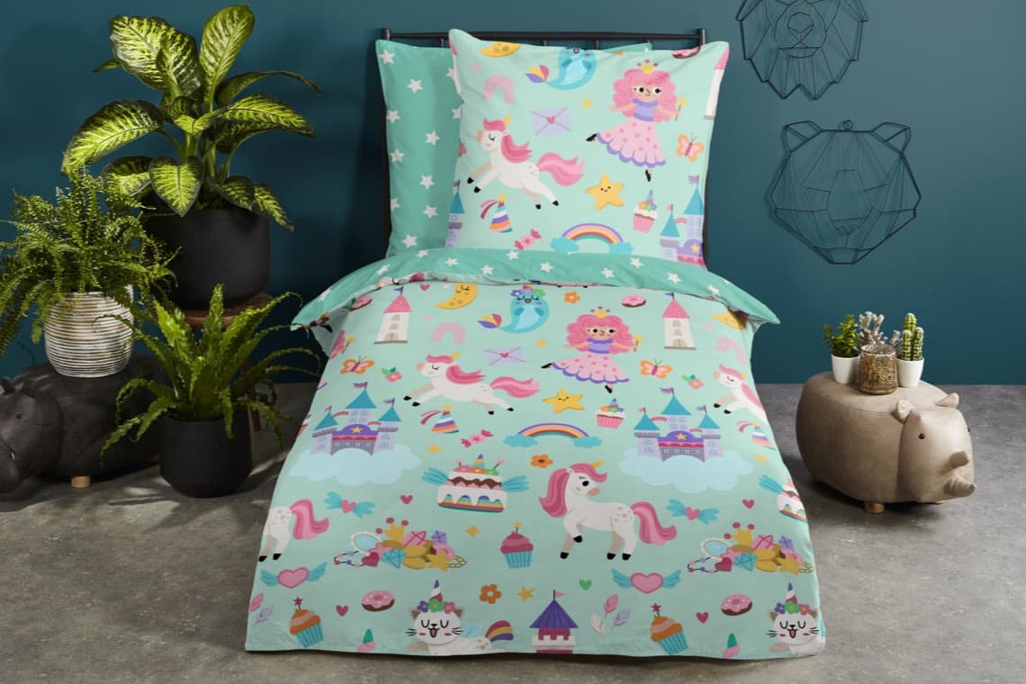 Kinderbett mit türkisfarbener Bettwäsche, bedruckt mit Einhörnern, Prinzessinnen, Regenbögen und Schlössern, umgeben von Pflanzen
