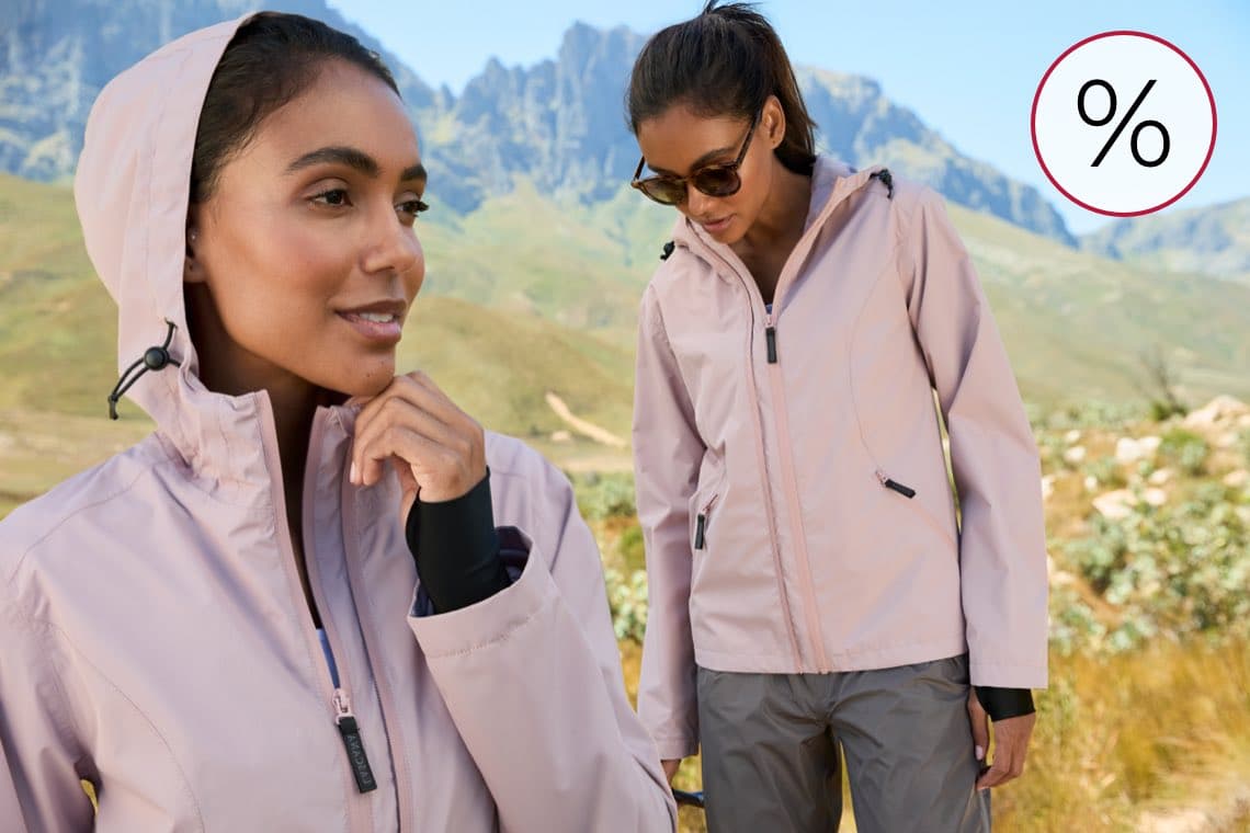 Prozentzeichen für Rabatt – Person mit hellrosafarbener Outdoorjacke wandert in bergiger Landschaft