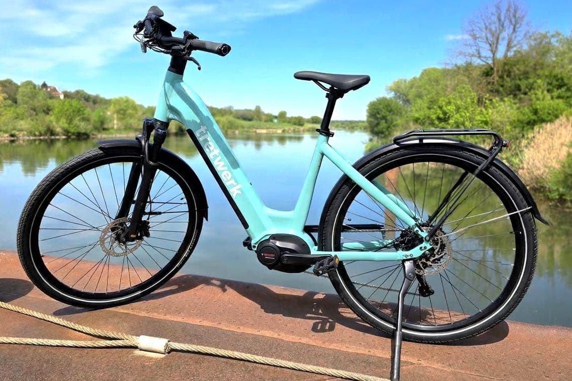 tretwerk E-Bike in Hellblau von der Seite, steht auf Steg vor ruhigem Fluss und grüner Landschaft