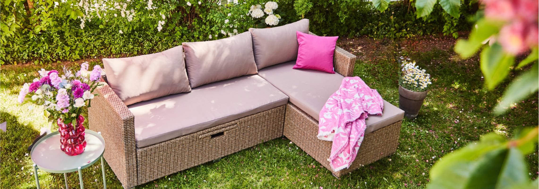 Rattan-Sofa im Garten mit rosa Kissen und Decke. Blumentopf und Tisch mit Blumen daneben.