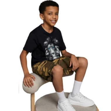 Junge sitzt auf Hocker, trägt schwarzes T‑Shirt mit Löwenmotiv, tarnfarbene Shorts und weiße Sneakers