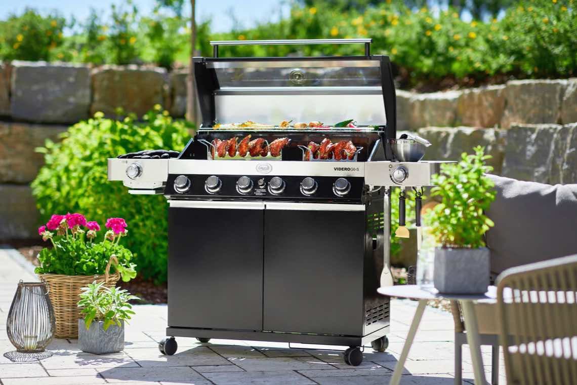 Großer Gasgrill im Garten, Grillgut auf dem Rost, umgeben von Pflanzen und Gartenmöbeln auf der Terrasse