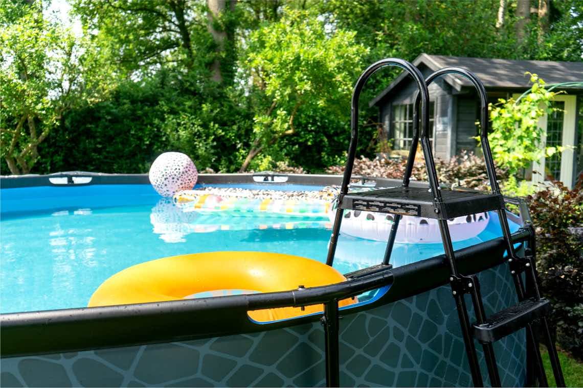 Aufstellpool im Garten mit Leiter, gelbem Schwimmring und bunten Luftmatratzen vor einem Gartenhaus