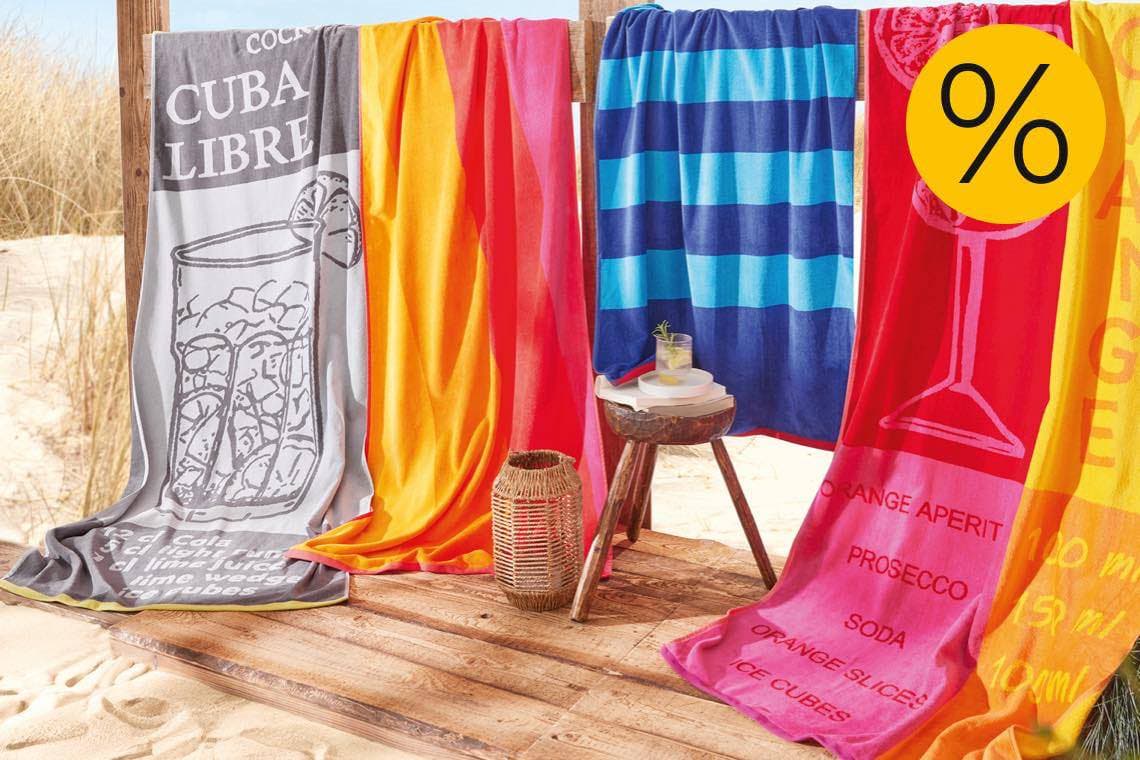Prozentzeichen für Rabatt – bunte Strandtücher mit Cocktailmotiven hängen an Holzgestell am Sandstrand