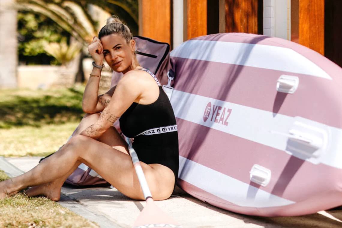 Frau im Badeanzug sitzt am Boden vor rosa YEAZ-SUP-Board und hält ein Paddel