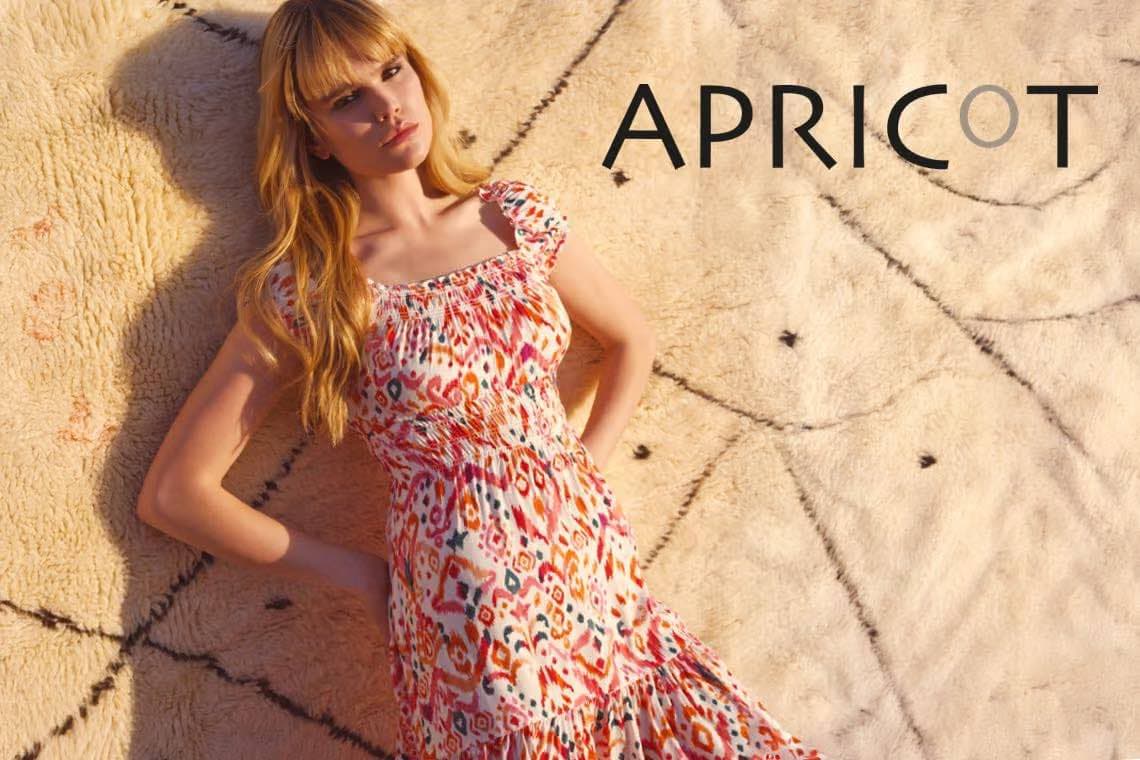 APRICOT – Frau in gemustertem Sommerkleid lehnt auf hellem Teppich und blickt in die Kamera