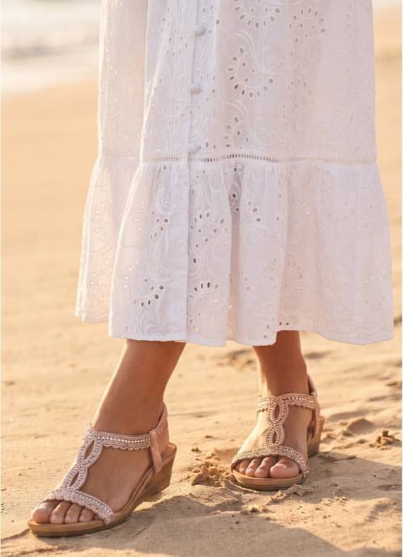 Frau in weißem Sommerkleid trägt verzierte Sandalen und geht am Strand entlang