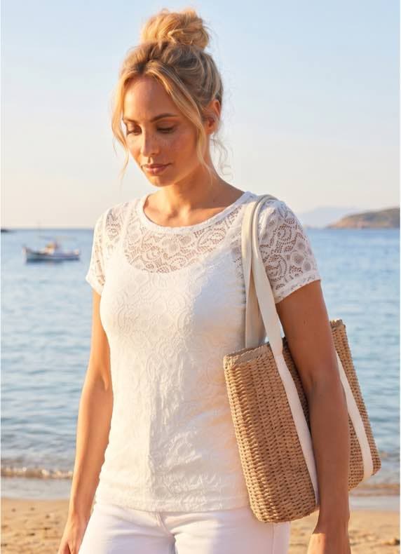 Frau im hellen Spitzen-T-Shirt mit großer Strandtasche geht am Meer entlang, Boote und Küste im Hintergrund