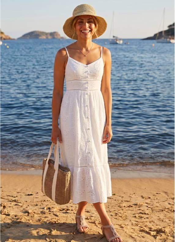 Frau im weißen Sommerkleid und Strohhut steht barfuß mit Basttasche am Strand vor ruhigem Meer