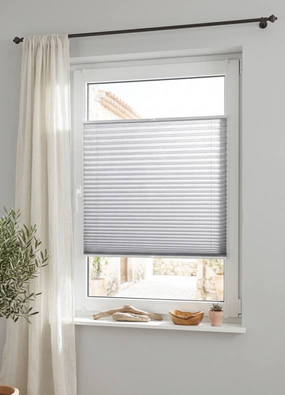 Graues Plissee am unteren Teil eines Fensters, daneben helle Gardine und Deko auf der Fensterbank