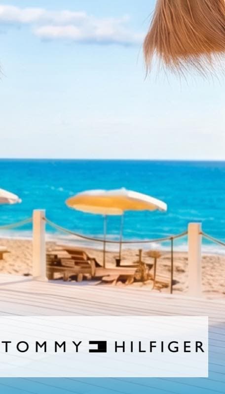 Bis 20.07. 22% auf TOMMY HILFIGER – Logo vor Holzterrasse mit Liegestühlen und Sonnenschirmen am Sandstrand mit blauem Meer