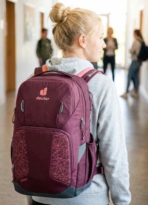 Person mit bordeauxfarbenem Deuter-Rucksack steht im Flur, im Hintergrund weitere Menschen in Gesprächen