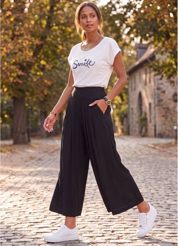 T-Shirt mit Aufdruck „Smile“ – Frau geht auf Kopfsteinpflaster, trägt weite schwarze Hose und weiße Sneaker
