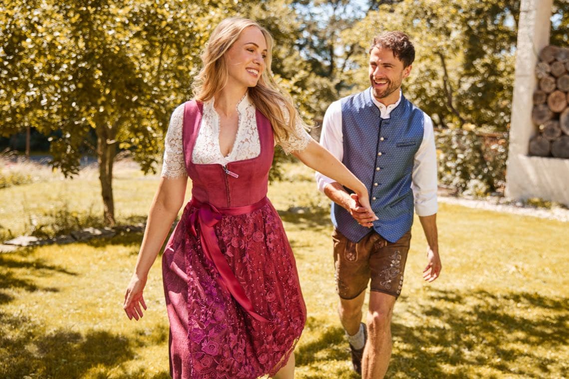 Frau im rosafarbenen Dirndl läuft über eine Wiese und hält die Hand eines Mannes in Trachtenweste und Lederhose