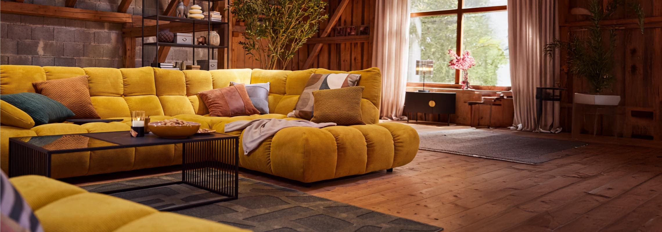 Großes gelbes Ecksofa mit bunten Kissen in rustikalem Wohnzimmer, davor schwarzer Couchtisch mit Snacks auf Holzboden