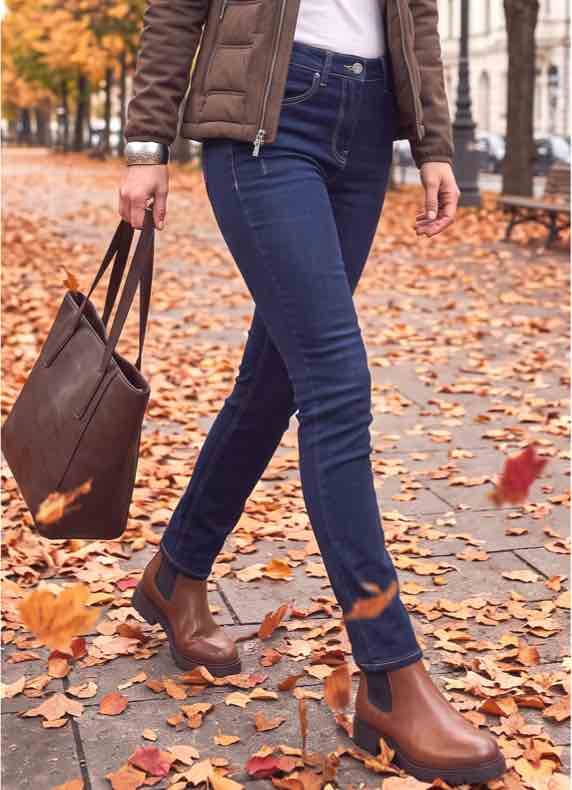 Frau in blauer Jeans und braunen Stiefeletten geht mit brauner Tasche über herbstliche Straße voller Laub