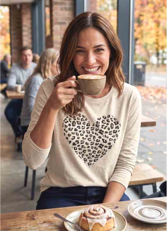 Frau im Café mit Cappuccino trägt beigen Pullover mit großem Herz in Leopardenmuster, Zimtschnecke auf dem Tisch