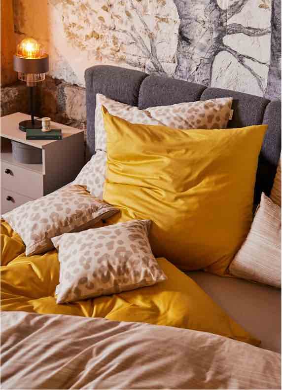 Bett mit gelber Bettwäsche und mehreren gemusterten Kissen vor grauem Kopfteil und Nachttisch mit Lampe