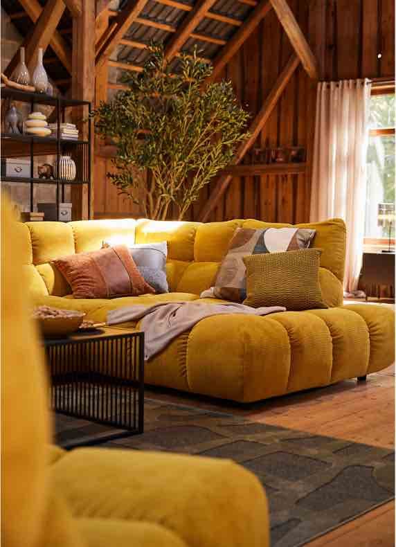 Gelbes Ecksofa mit bunten Kissen und Decke in rustikalem Wohnzimmer mit Holzdecke, Regal und großem Fenster