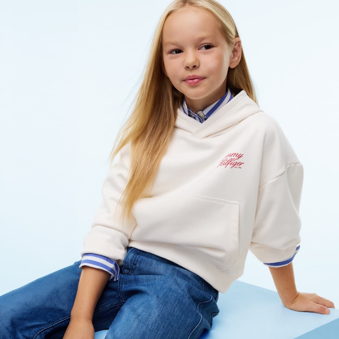 Mädchen mit langen Haaren sitzt in hellem Hoodie und Jeans vor hellblauem Hintergrund