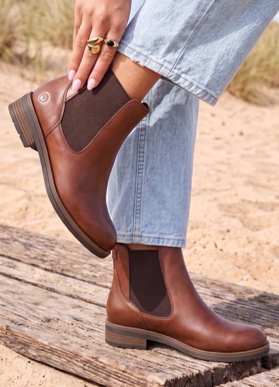 Person in hellbrauner Jeans trägt braune Stiefeletten am Strand und streicht mit der Hand über den Schaft
