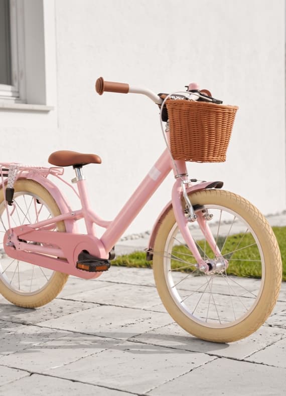Rosafarbenes Kinderfahrrad mit Korb am Lenker steht auf gepflasterter Terrasse vor weißer Hauswand