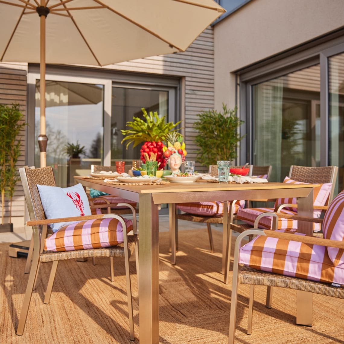 Terrassentisch mit vier Stühlen, gestreiften Sitzkissen und Geschirr unter großem Sonnenschirm, umgeben von Pflanzen